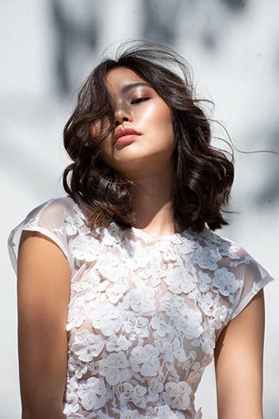 Braut in einem zarten Spitzenkleid mit floralem Muster und welligem Haar. Ihr Gesicht ist entspannt, während sie das Licht auf ihrer Haut genießt, was eine ruhige Atmosphäre schafft.