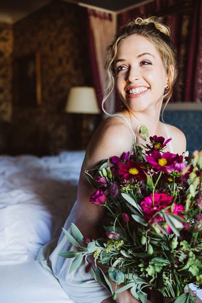 Braut in einem eleganten, weißen Kleid sitzt auf einem Bett mit einem bunten Blumenstrauß in der Hand. Sie lächelt glücklich und wirkt entspannt.