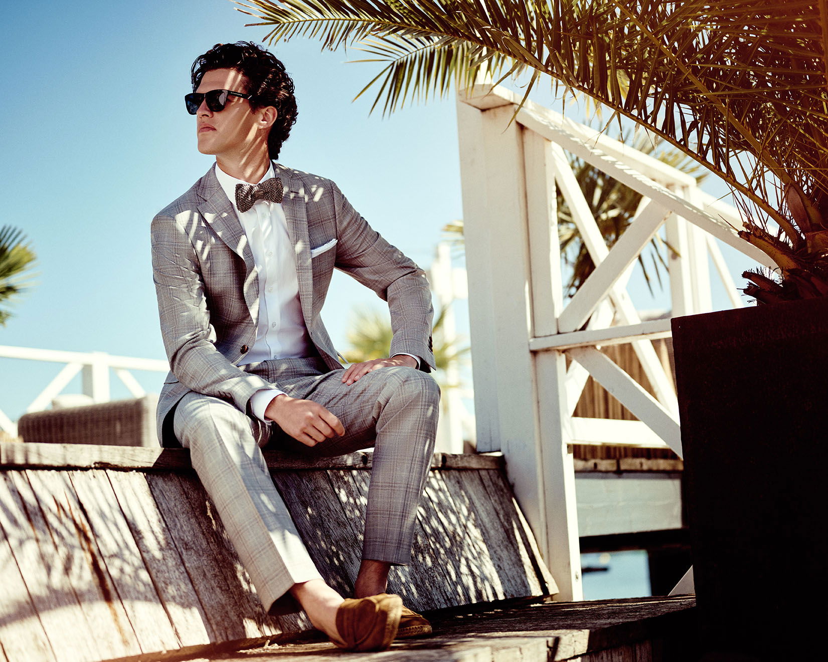 Stilvoller Mann in einem maßgeschneiderten grauen Anzug und einer Fliege entspannt sich unter Palmen, während die Sonne ihn an der Küste anstrahlt.