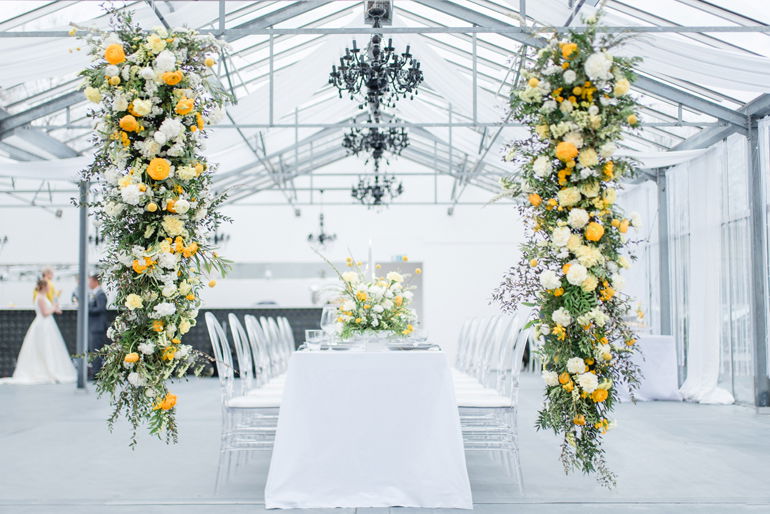 Ein festlich gedeckter Tisch in einem modernen Gewächshaus, flankiert von hohen Blumenarrangements in Gelb und Weiß, unter schwebenden Kronleuchtern.