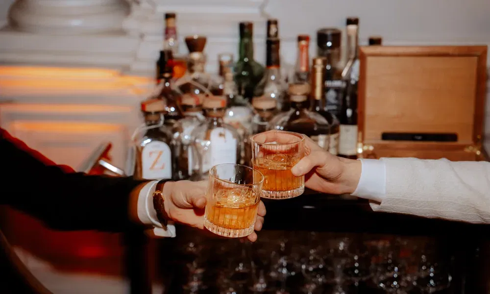Zwei Männer stoßen mit Drinks an. Es sidn ur ihre Arme zu sehen. Dahinter steht ein Barwagen.
