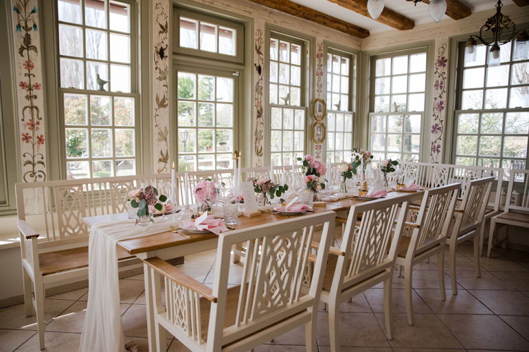 Hell und romantisch dekorierter Hochzeitstisch mit Blumenarrangements in einem Raum mit vielen Fenstern und eleganten weißen Holzmöbeln.