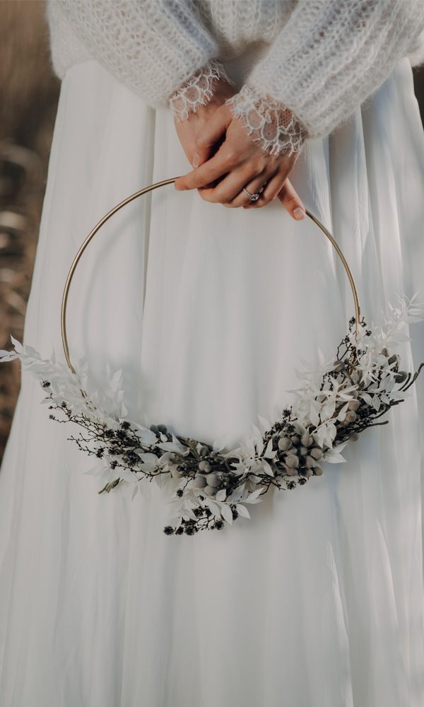 Detailaufnahme einer Braut in einem romantischen Strickoberteil mit Spitzenbündchen, die einen minimalistischen goldenen Hoop mit weißen Trockenblumen und dunklen Akzenten hält.