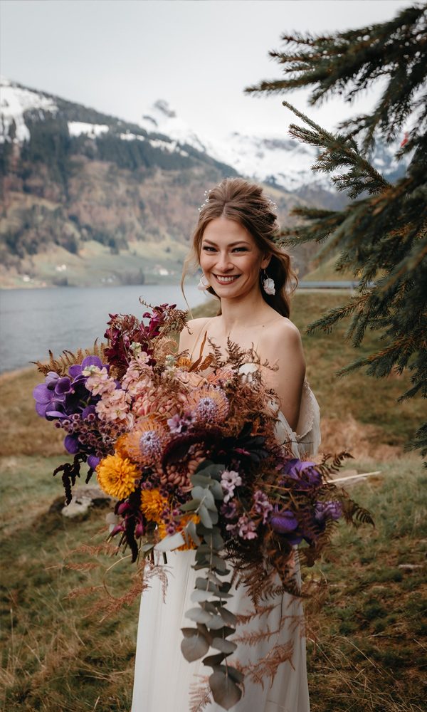 Eine fröhliche Braut mit einem beeindruckenden, wilden Blumenstrauß aus herbstlichen und violetten Tönen steht in einer malerischen Landschaft mit schneebedeckten Bergen und einem See.