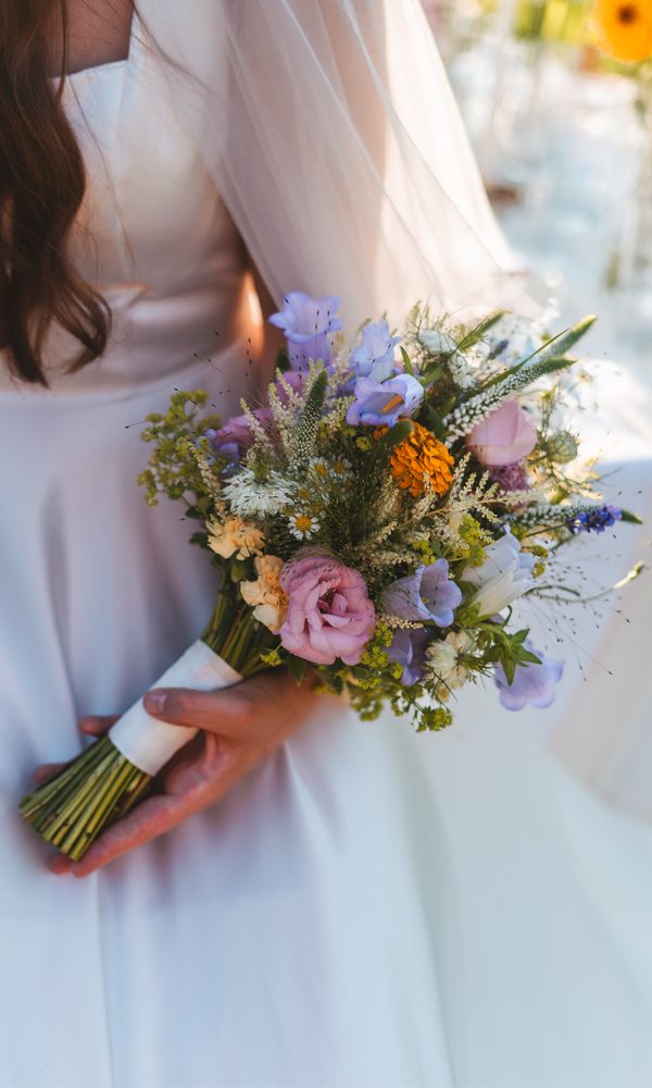 Nahaufnahme einer Braut mit einem romantischen, locker gebundenen Hochzeitsstrauß aus bunten Wiesenblumen. Ihr weißes Kleid aus glänzendem Stoff reflektiert das warme Sonnenlicht.