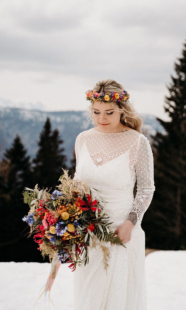 Eine winterliche Braut mit offenen, blonden Haaren trägt ein weißes Spitzenkleid mit transparenten Ärmeln und einen leuchtenden Blumenkranz. Ihr großer Hochzeitsstrauß kombiniert getrocknete und frische Blüten in kräftigen Farben.