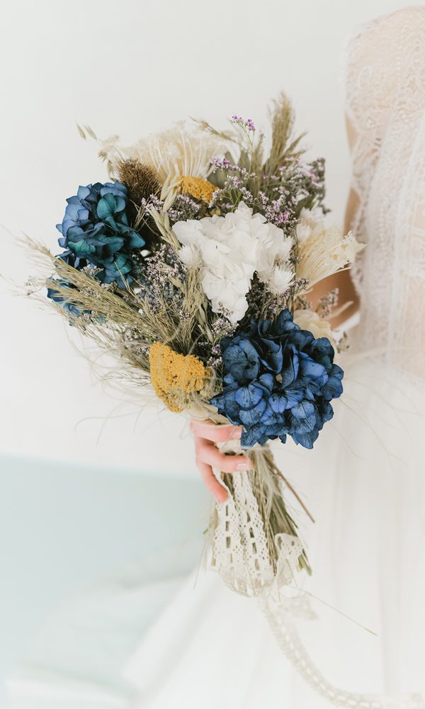 Detailaufnahme eines kunstvollen Brautstraußes mit leuchtend blauen Hortensien, weißen Blüten und getrockneten Gräsern. Die Braut trägt ein romantisches Kleid mit feiner Spitze.