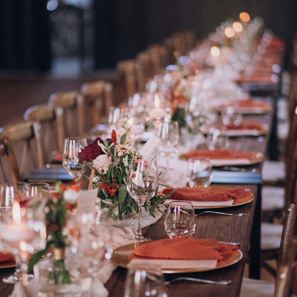 Ein festlich dekorierter Hochzeitstisch, auf dem Gläser, Besteck und orangefarbene Servietten perfekt arrangiert sind. Blumenarrangements und Kerzenlicht sorgen für eine warme Atmosphäre.