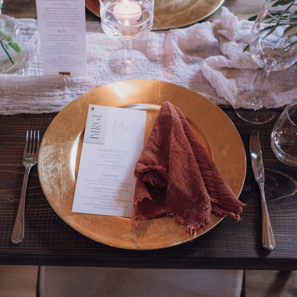 Nahaufnahme eines Hochzeitstisches mit einem goldenen Teller, einer orangefarbenen Stoffserviette und einem Menü. Eine Kerze im Glas und eine elegante Blumenvase mit roten Akzenten schmücken den Tisch.