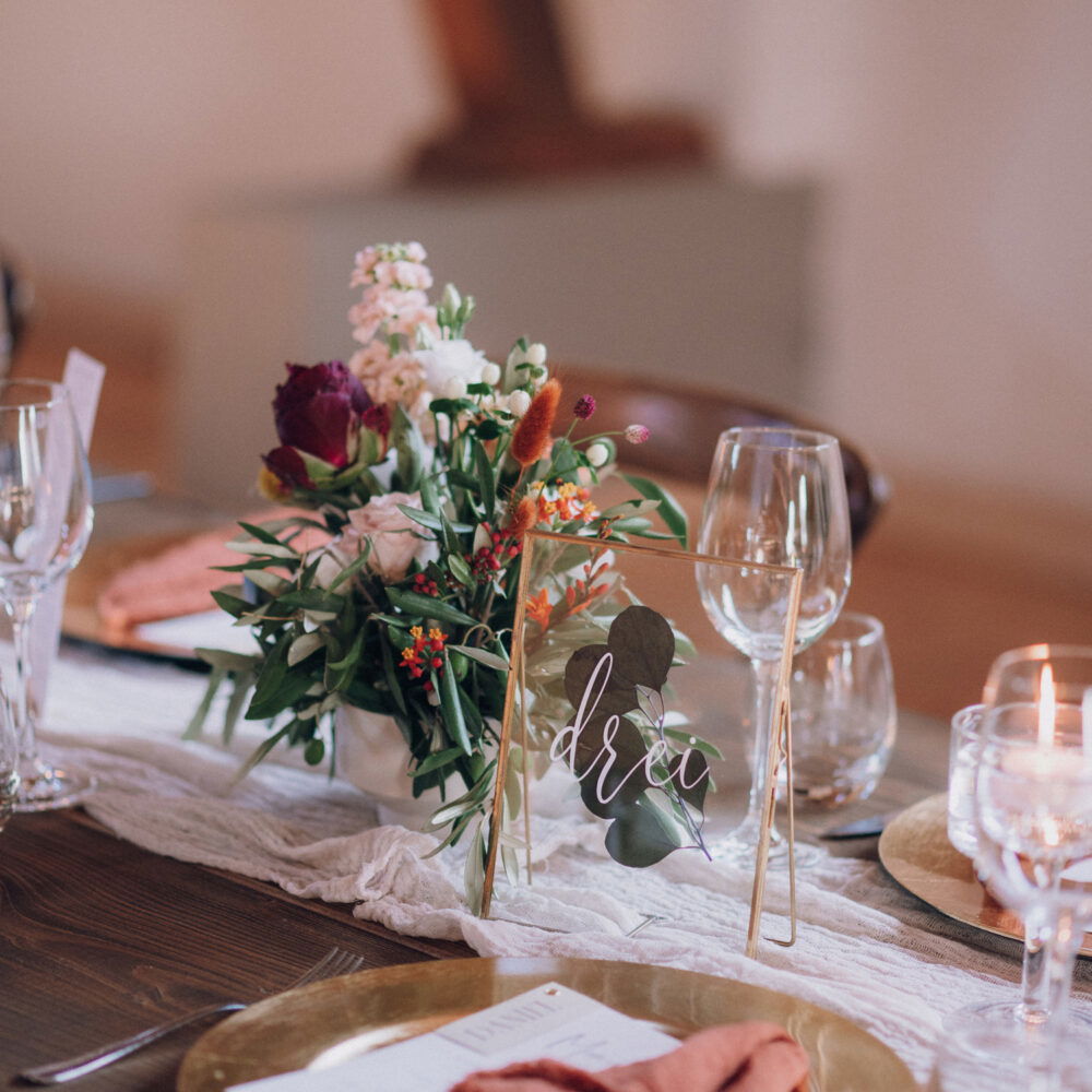 Ein festlich gedeckter Tisch mit Blumenarrangements und einem Namensschild „Drei“. Auf dem Tisch stehen Weingläser, goldene Teller und dunkelrote Servietten. Ein leuchtendes Kerzenlicht sorgt für eine romantische Atmosphäre.