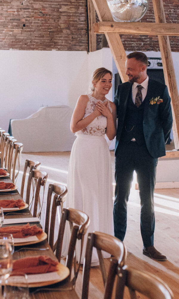 Ein emotionaler Moment: Die Braut und der Bräutigam bestaunen ihren festlich geschmückten Hochzeitssaal. Der rustikale Charme des Raums mit Holzbalken, Kerzen und liebevoll gestalteter Tischdekoration sorgt für eine romantische Stimmung.
