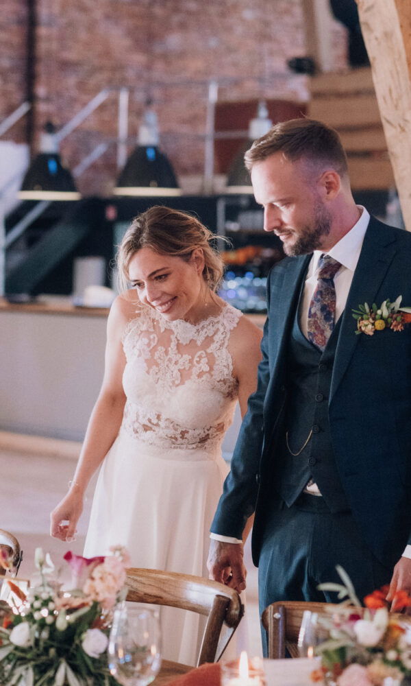 Das Brautpaar betrachtet die festlich gedeckte Hochzeitstafel mit leuchtenden Augen. Die Braut strahlt vor Freude, während der Bräutigam sich die Dekoration genau ansieht. Der rustikale Raum mit Holzbalken verleiht der Szene eine besondere Note.