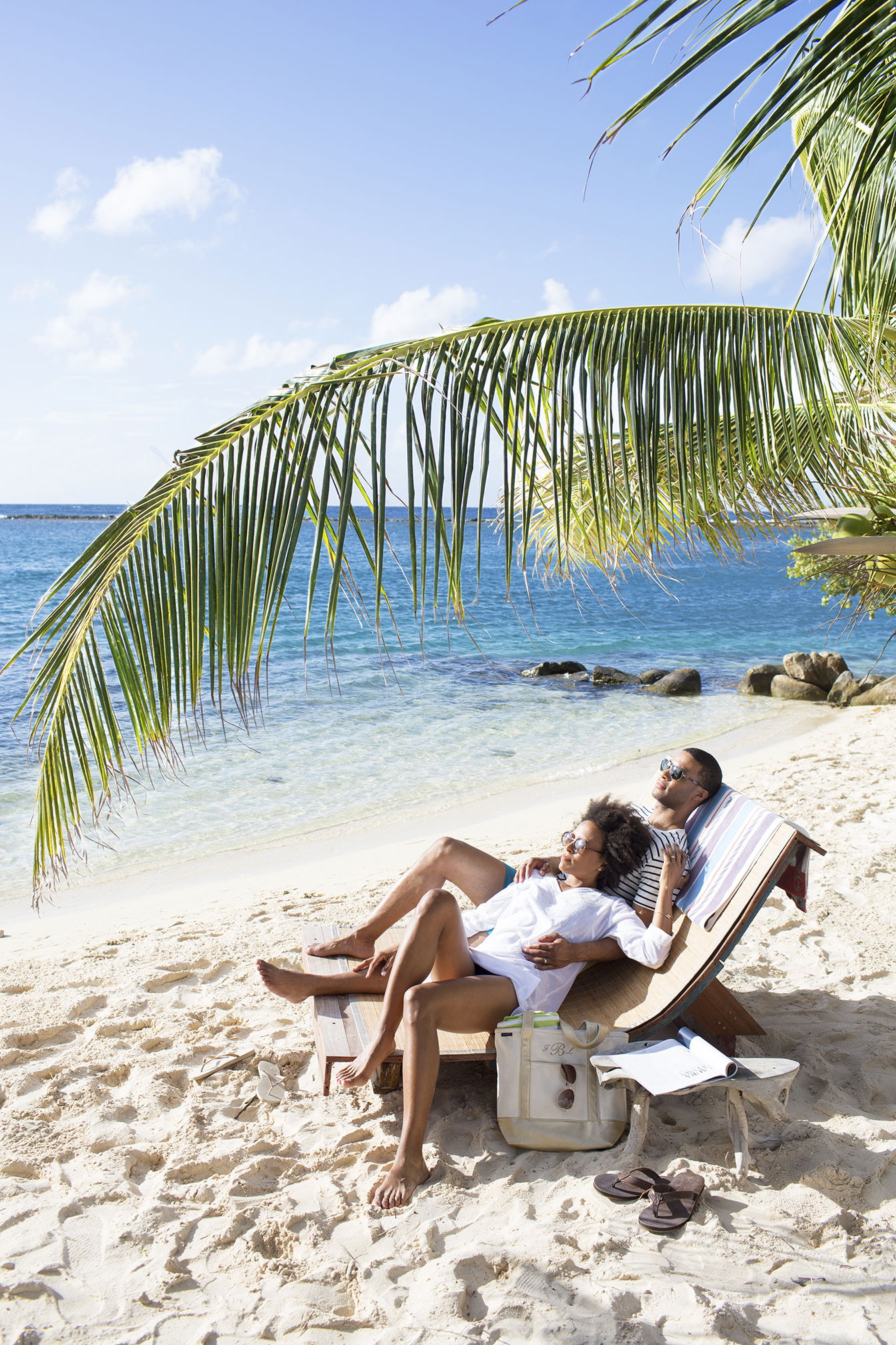 Ein Paar genießt die Flitterwochen am Strand, entspannt auf einer Holzliege unter einer Palme. Sie blicken auf das ruhige Meer, während eine Strandtasche und Sandalen im Vordergrund im Sand liegen.