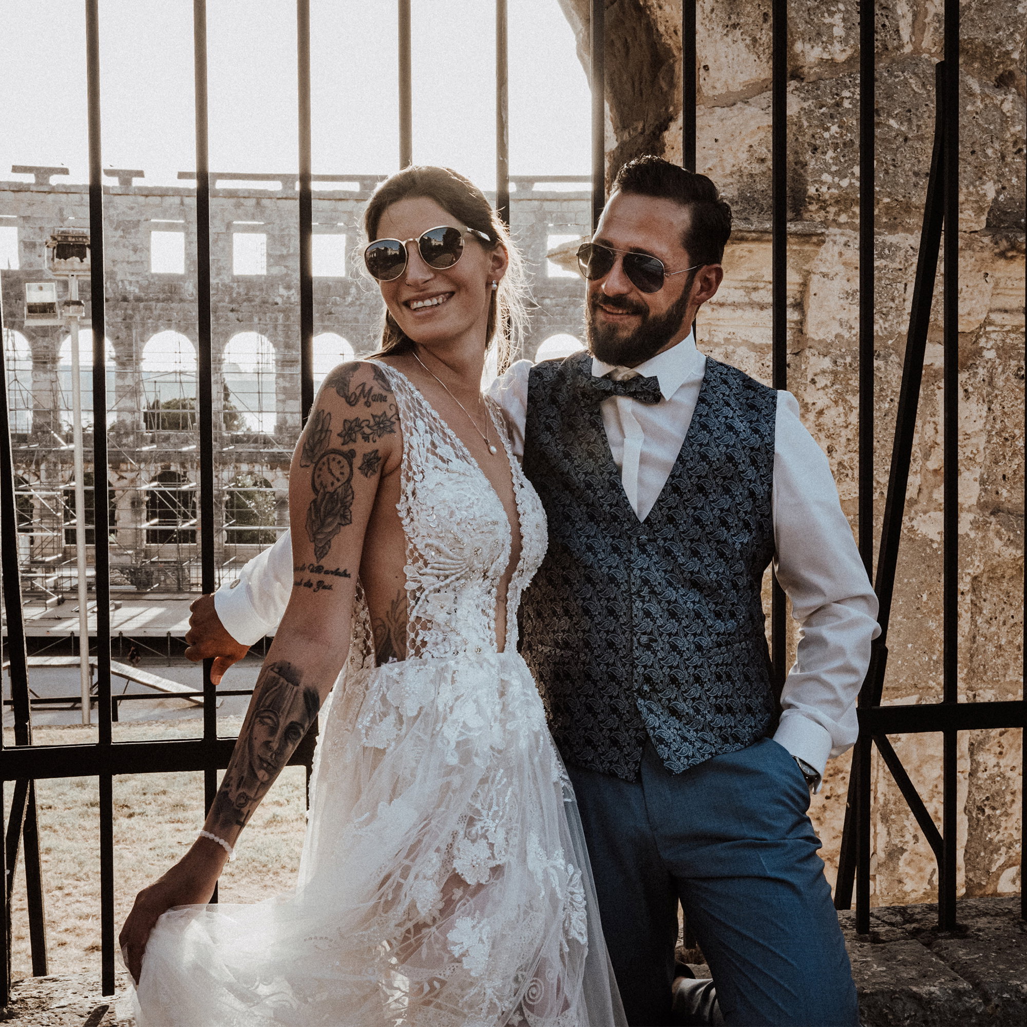 Das Brautpaar genießt den Moment in Sonnenbrillen und Hochzeitskleidung, lächelnd vor einer antiken Kulisse.