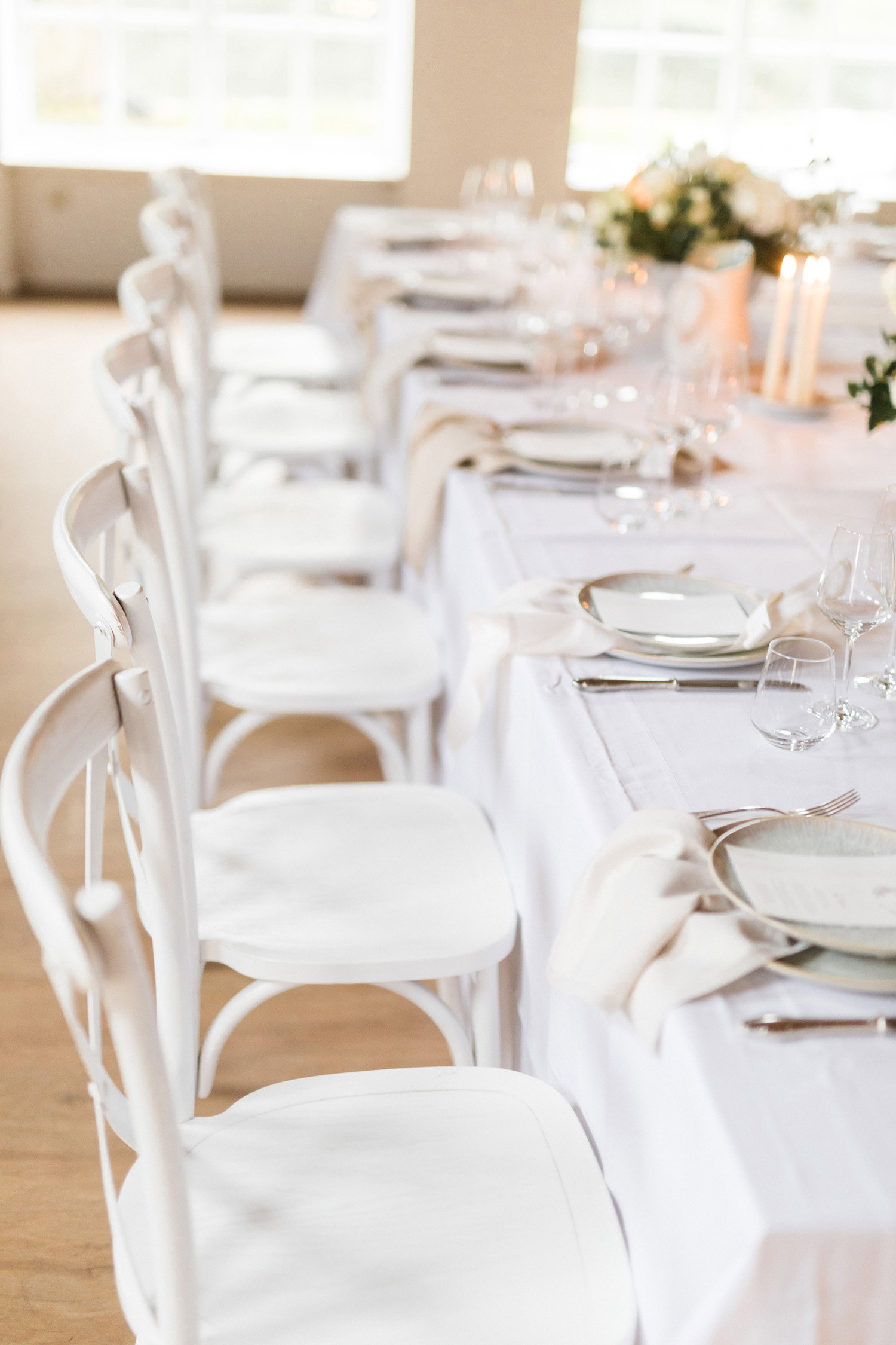 Schlichte und elegante Hochzeitstafel in Weißtönen, mit ordentlich angeordneten Stühlen und einer langen Tafel, bereit für die Gäste.