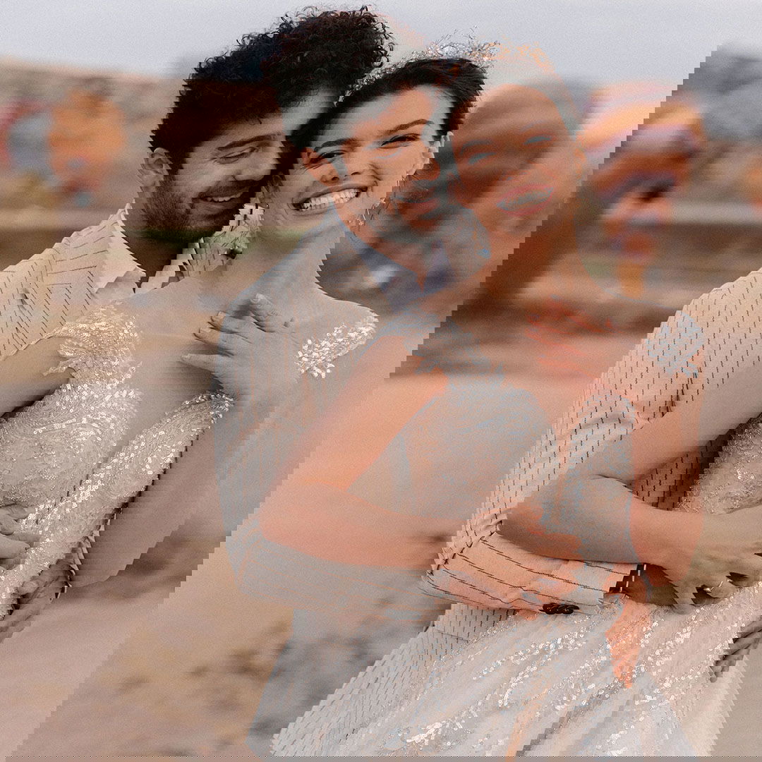 Braut und Bräutigam lachen gemeinsam in die Kamera, umgeben von einer malerischen Landschaft mit Heißluftballons im Hintergrund.