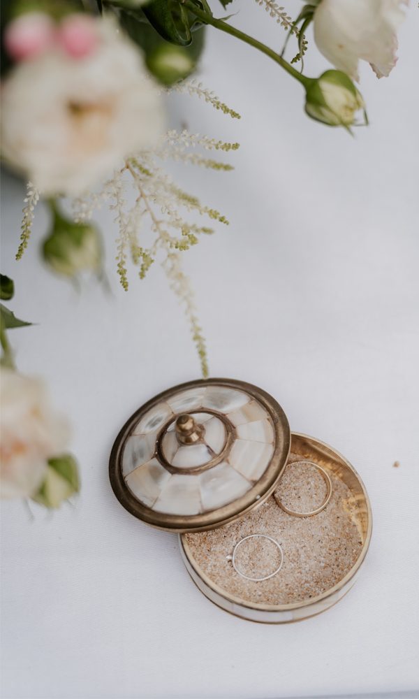 Eine geöffnete Schmuckdose mit zwei zarten Eheringen, eingebettet in Sand. Die Szene wird von weißen Blumen umrahmt und erzeugt eine natürliche, elegante Hochzeitsatmosphäre.