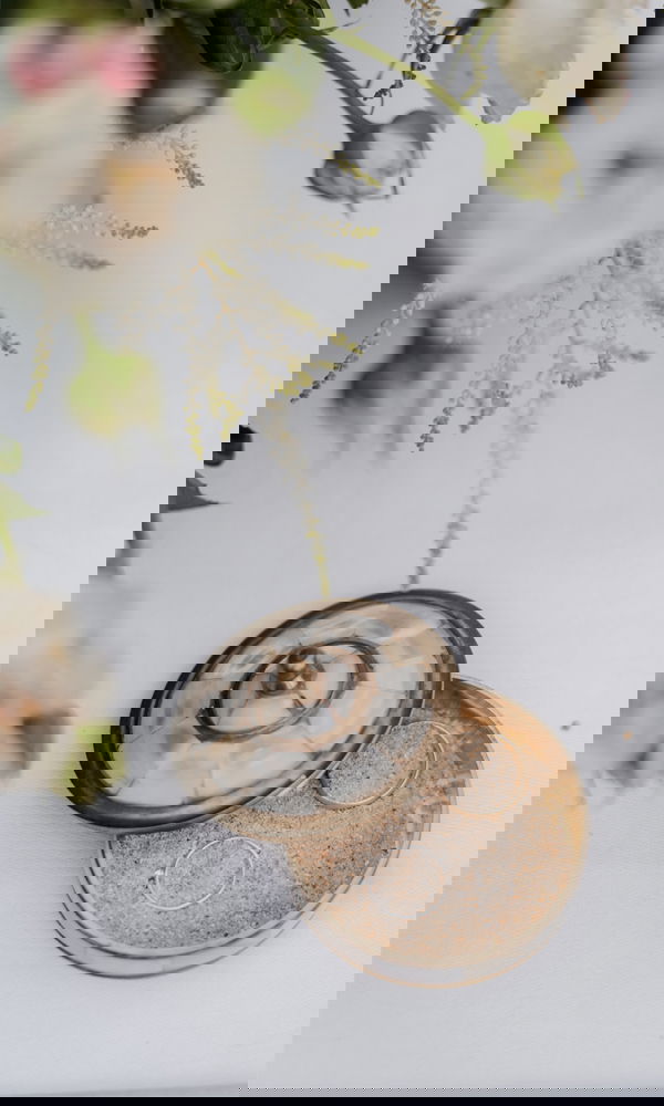 Eine geöffnete Schmuckdose mit zwei zarten Eheringen, eingebettet in Sand. Die Szene wird von weißen Blumen umrahmt und erzeugt eine natürliche, elegante Hochzeitsatmosphäre.