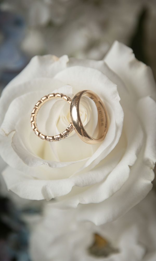 Zwei Hochzeitsringe in einer frischen, weißen Blüte arrangiert. Der eine Ring hat eine glatte, polierte Oberfläche, der andere besticht durch eine feine Struktur.