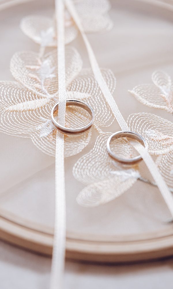 Minimalistische Hochzeitsringe, die auf einem transparenten Stickrahmen mit feinen Blumenstickereien liegen. Die cremefarbenen Bänder halten die Ringe sicher an ihrem Platz.
