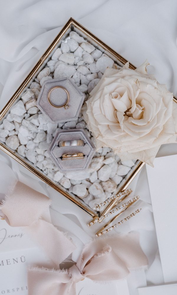 Hochzeitsringe in einer eleganten, hexagonalen Schmuckbox, kunstvoll arrangiert auf weißen Steinen. Die Szene wird durch zarte Stoffbänder, eine cremefarbene Rose und goldene Haaraccessoires ergänzt.