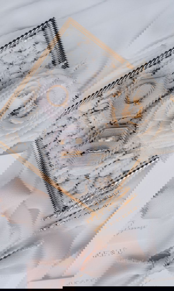 Hochzeitsringe in einer eleganten, hexagonalen Schmuckbox, kunstvoll arrangiert auf weißen Steinen. Die Szene wird durch zarte Stoffbänder, eine cremefarbene Rose und goldene Haaraccessoires ergänzt.