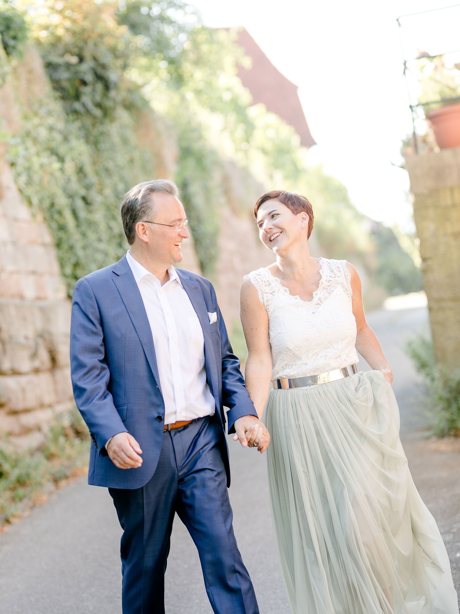 Ein Paar geht lachend nebeneinander her und hält sich an den Händen. Der Mann trägt einen klassischen blauen Anzug, während die Frau ein weißes Spitzenoberteil und einen langen, fließenden Rock trägt. Der Weg ist von Pflanzen und alten Mauern umgeben.