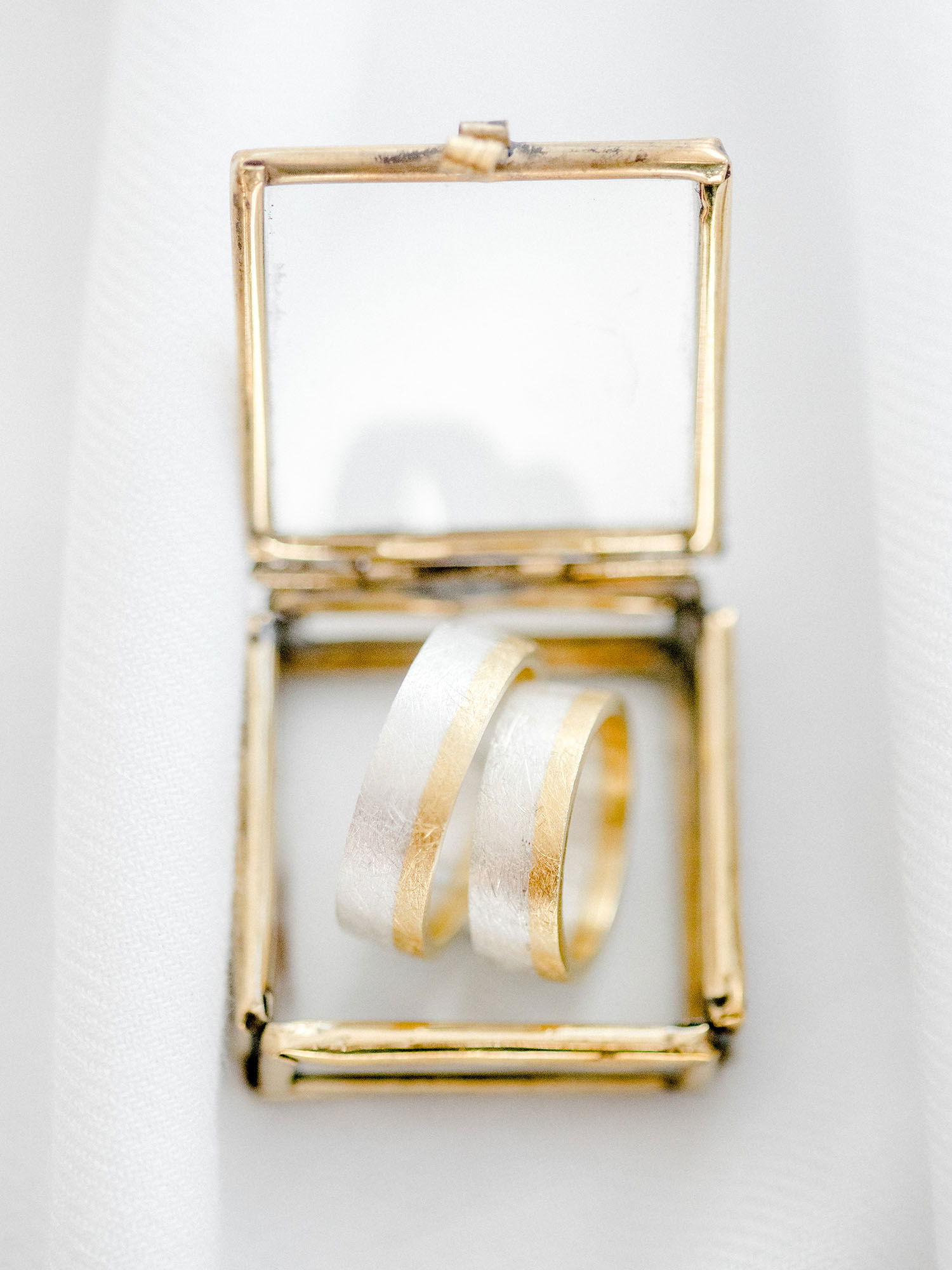 Detailaufnahme von zwei eleganten Eheringen in einer kleinen, quadratischen Schatulle aus goldfarbenem Metall. Die Ringe haben einen breiten, mattierten Streifen, der von einem glänzenden Rand eingerahmt ist.