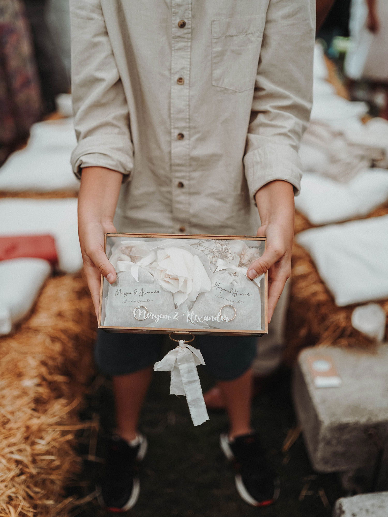 Ein Kind präsentiert eine dekorative Box mit den Namen des Hochzeitspaares, Meryem und Alexander, verziert mit weißen Stoffbändern. Die Szene spielt in einer rustikalen Umgebung mit Heuballen im Hintergrund.