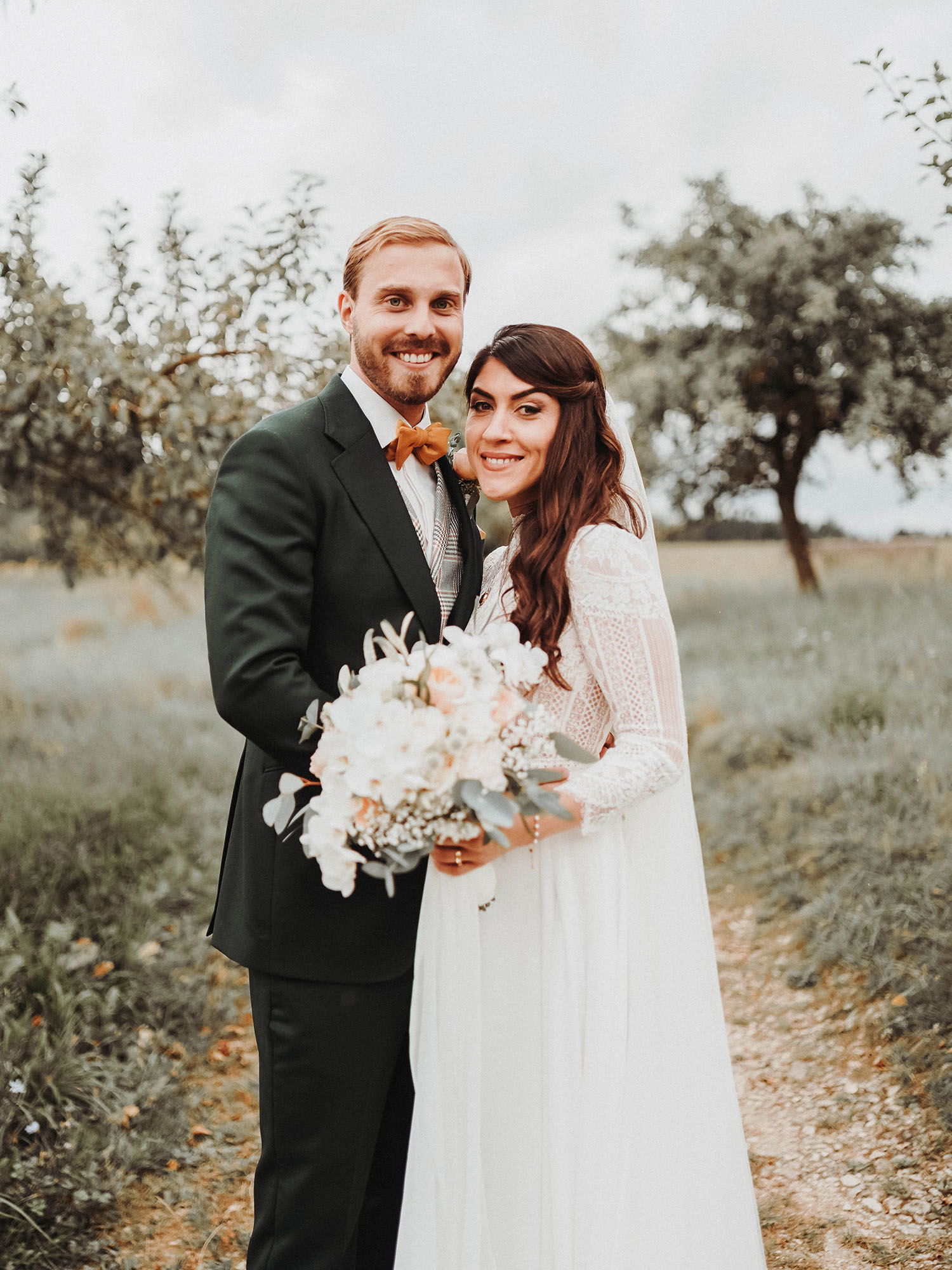 Meryem und Alexander stehen glücklich auf einem Pfad im Grünen. Der Bräutigam trägt einen grünen Anzug mit einer auffälligen orangefarbenen Fliege, und die Braut hält einen großen Blumenstrauß mit weißen und rosa Blumen.