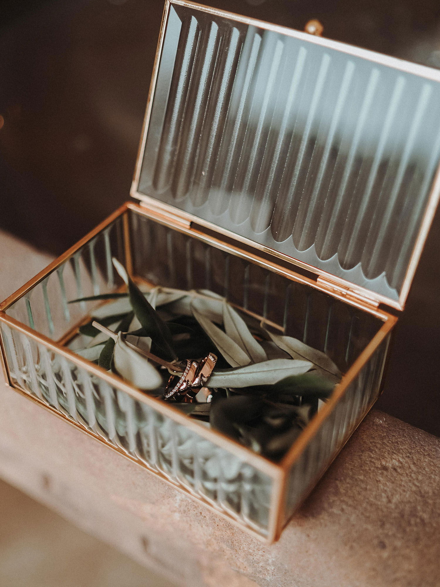 Detailaufnahme einer goldgerahmten Glasbox, die mit Olivenblättern und einem Paar Eheringen gefüllt ist. Die Box steht auf einem Steintisch und reflektiert sanft das Licht der Umgebung.