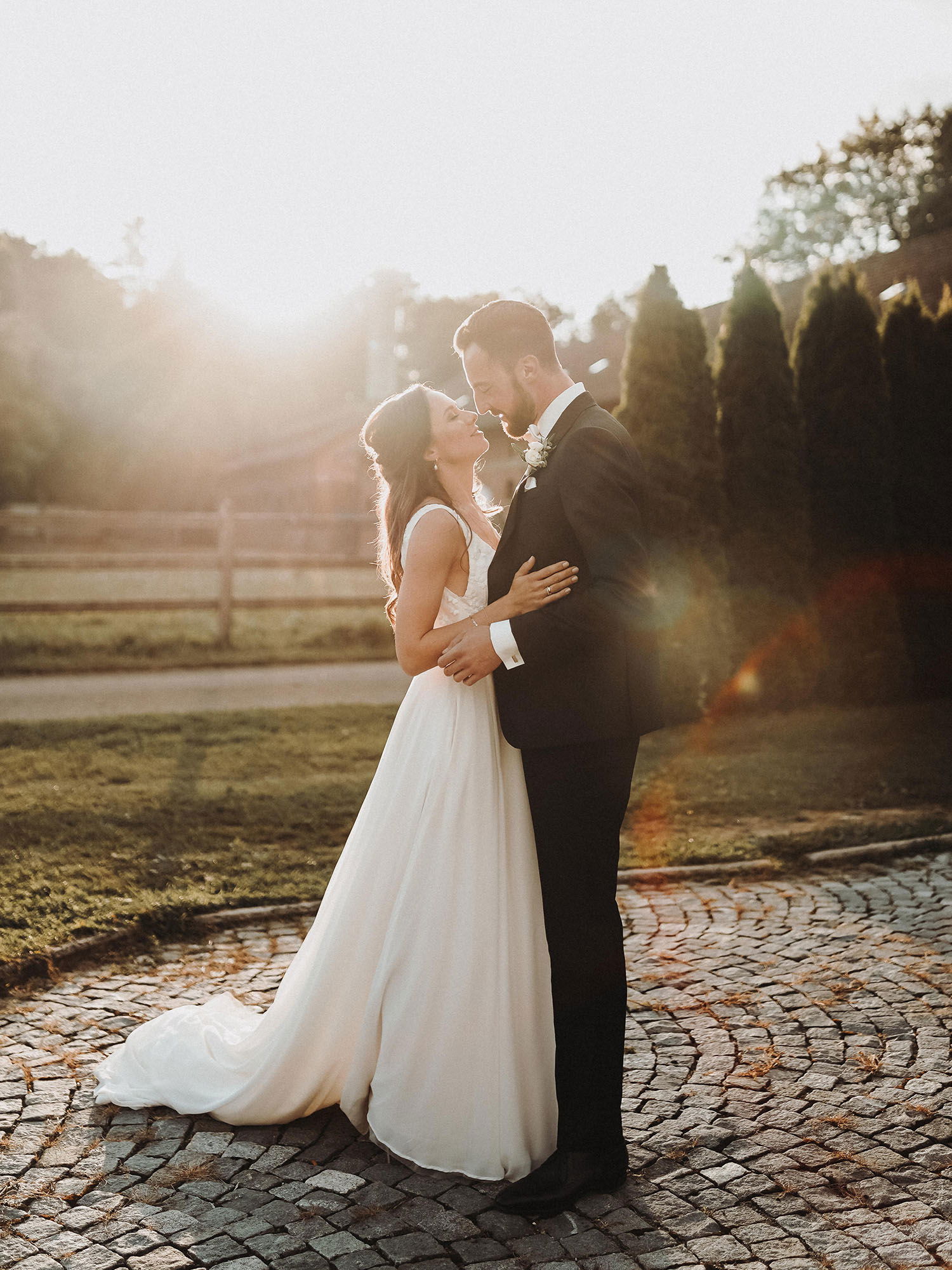 Ein Brautpaar umarmt sich zärtlich bei Sonnenuntergang auf einem gepflasterten Weg. Die Braut trägt ein schlichtes, aber elegantes weißes Kleid, während der Bräutigam einen schwarzen Anzug und eine Blumenanstecknadel trägt. Die Atmosphäre ist romantisch, unterstrichen vom warmen Licht der Abendsonne.