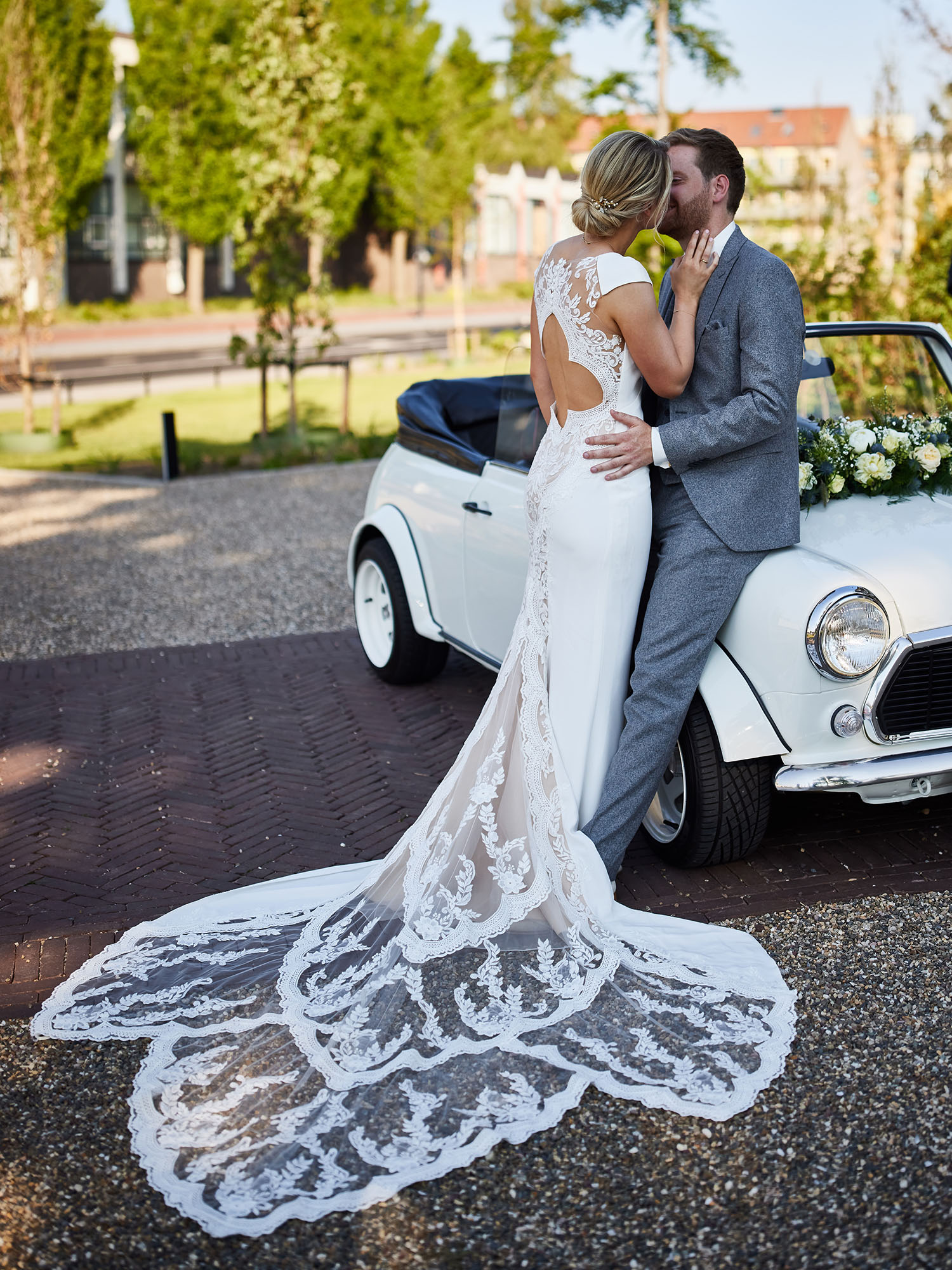 Ein frisch verheiratetes Paar steht vor einem klassischen, weißen Cabrio. Die Braut in einem Spitzenkleid mit langer Schleppe umarmt und küsst ihren Bräutigam, der sich lässig gegen das Auto lehnt. Die ländliche Umgebung und das Auto schaffen ein charmantes, romantisches Bild.