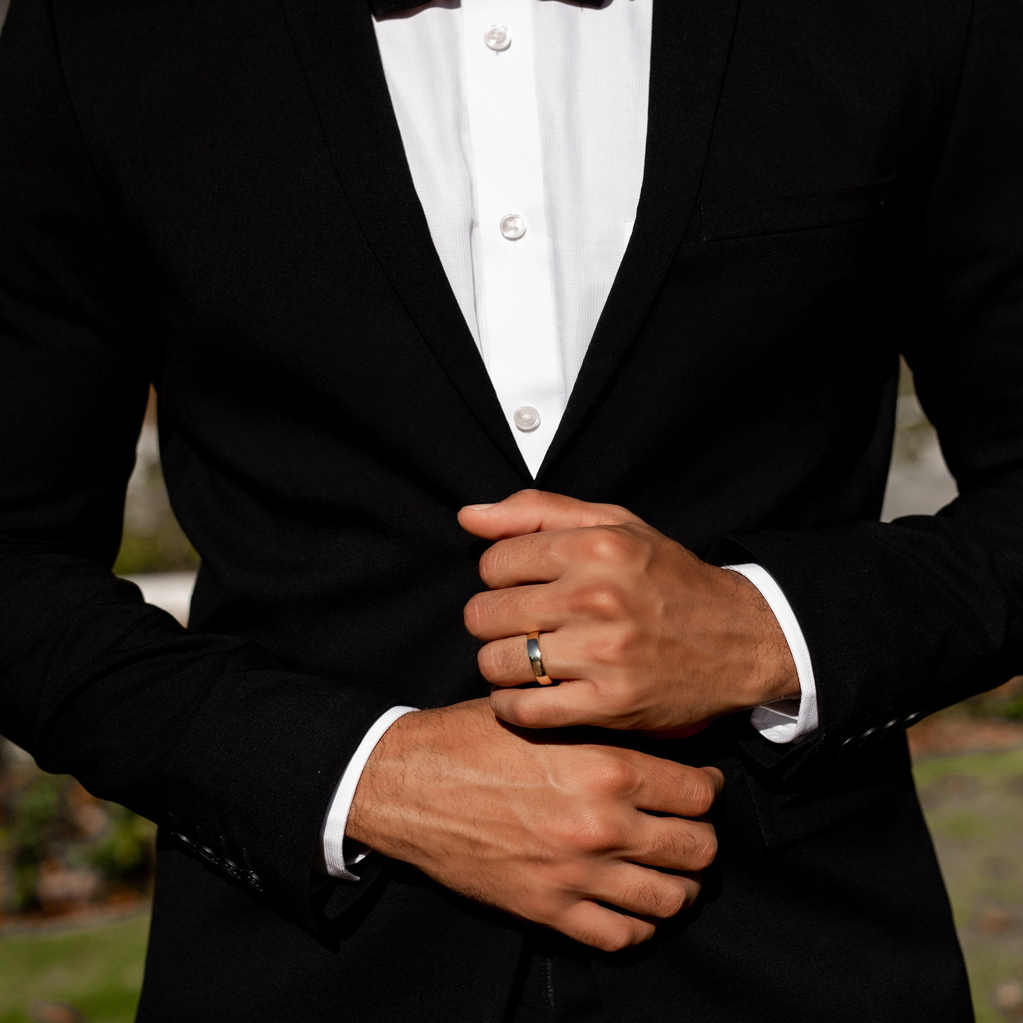 Der Bräutigam in einem stilvollen schwarzen Anzug und weißem Hemd hält seine Hände vor sich zusammen. Sein Ehering glänzt dezent an seiner Hand.