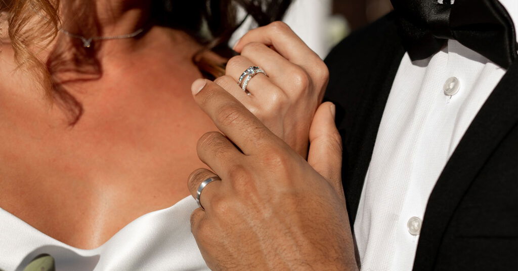 Das Brautpaar steht dicht beieinander, während sie sich liebevoll berühren. Ihre Hände und Ringe stehen im Fokus und symbolisieren ihre Verbundenheit.