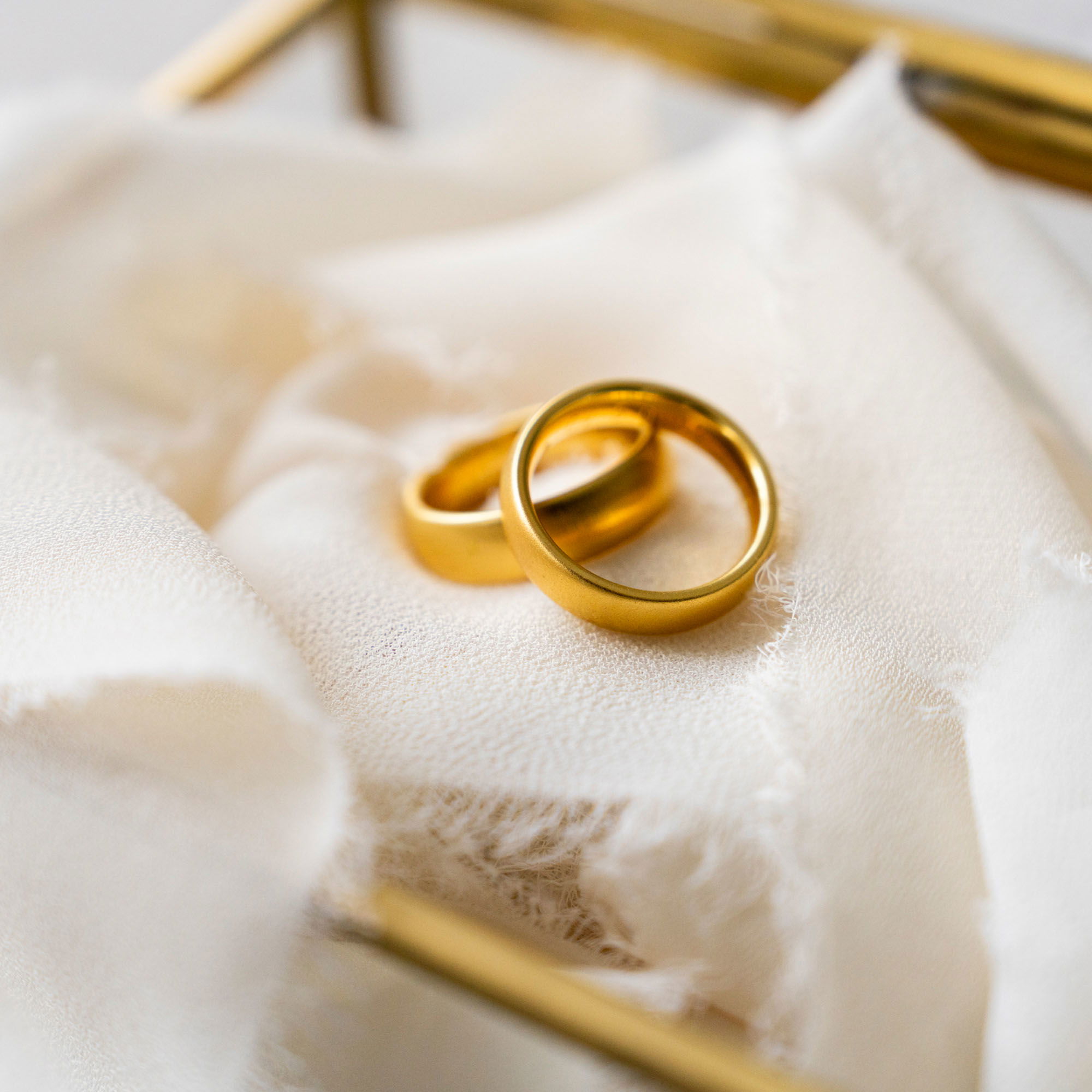 Nahaufnahme von zwei goldenen Eheringen, die auf zartem, beigem Stoff in einer dekorativen Glasbox präsentiert werden. Ein elegantes Arrangement für den Hochzeitstag.