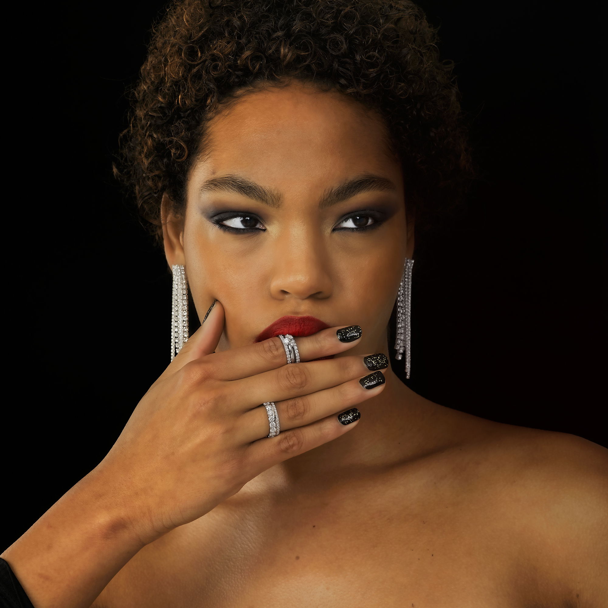 Eine Frau mit glamourösen Ohrringen und rauchigem Augen-Make-up trägt zwei funkelnde Ringe. Eine Hand mit dunklem Nagellack ruht auf ihrem Gesicht und betont die eleganten Schmuckstücke.
