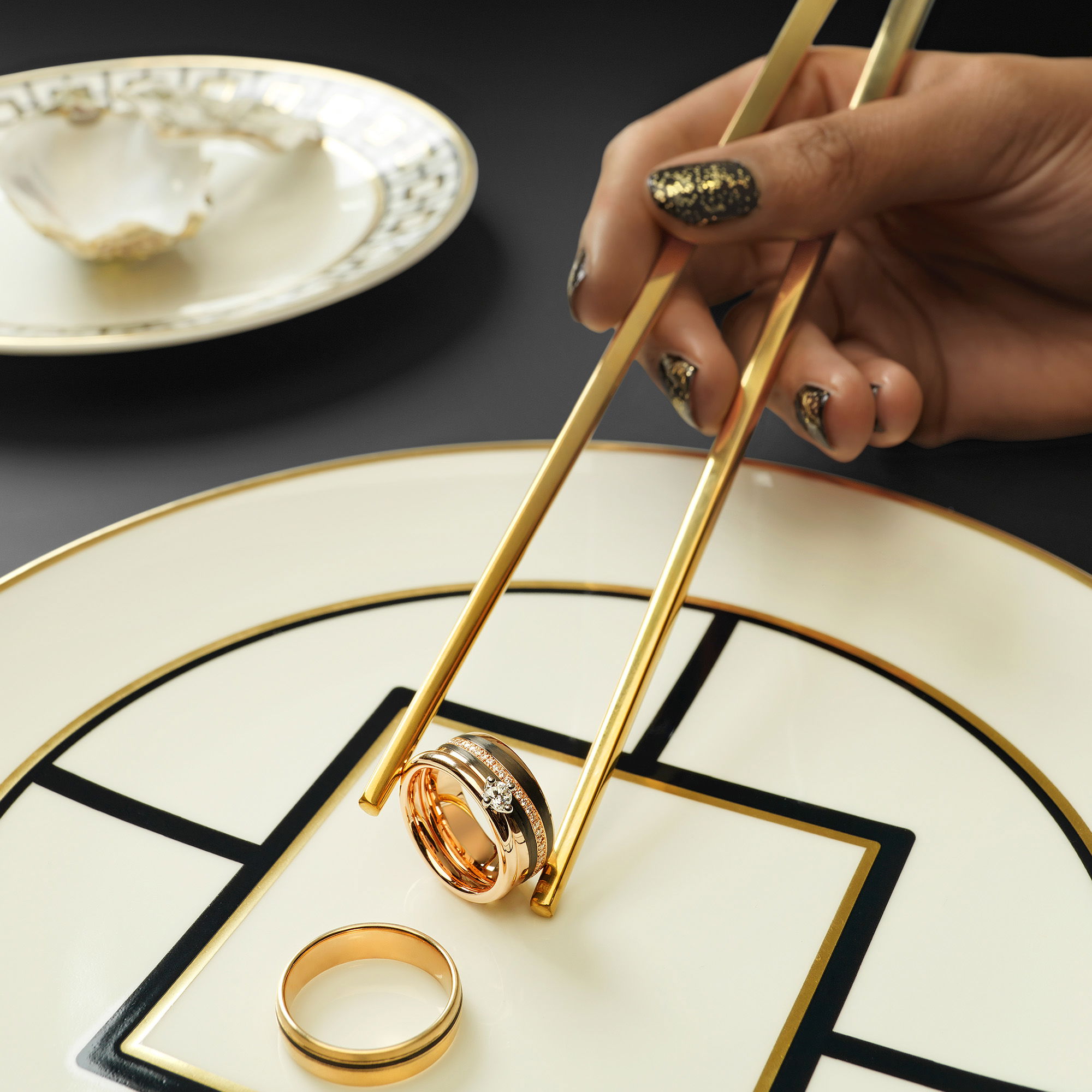 Auf einem kunstvollen Teller wird ein goldener Ehering mit Essstäbchen präsentiert, während ein zweiter Ehering daneben liegt. Ein originelles Arrangement für Trauringe.