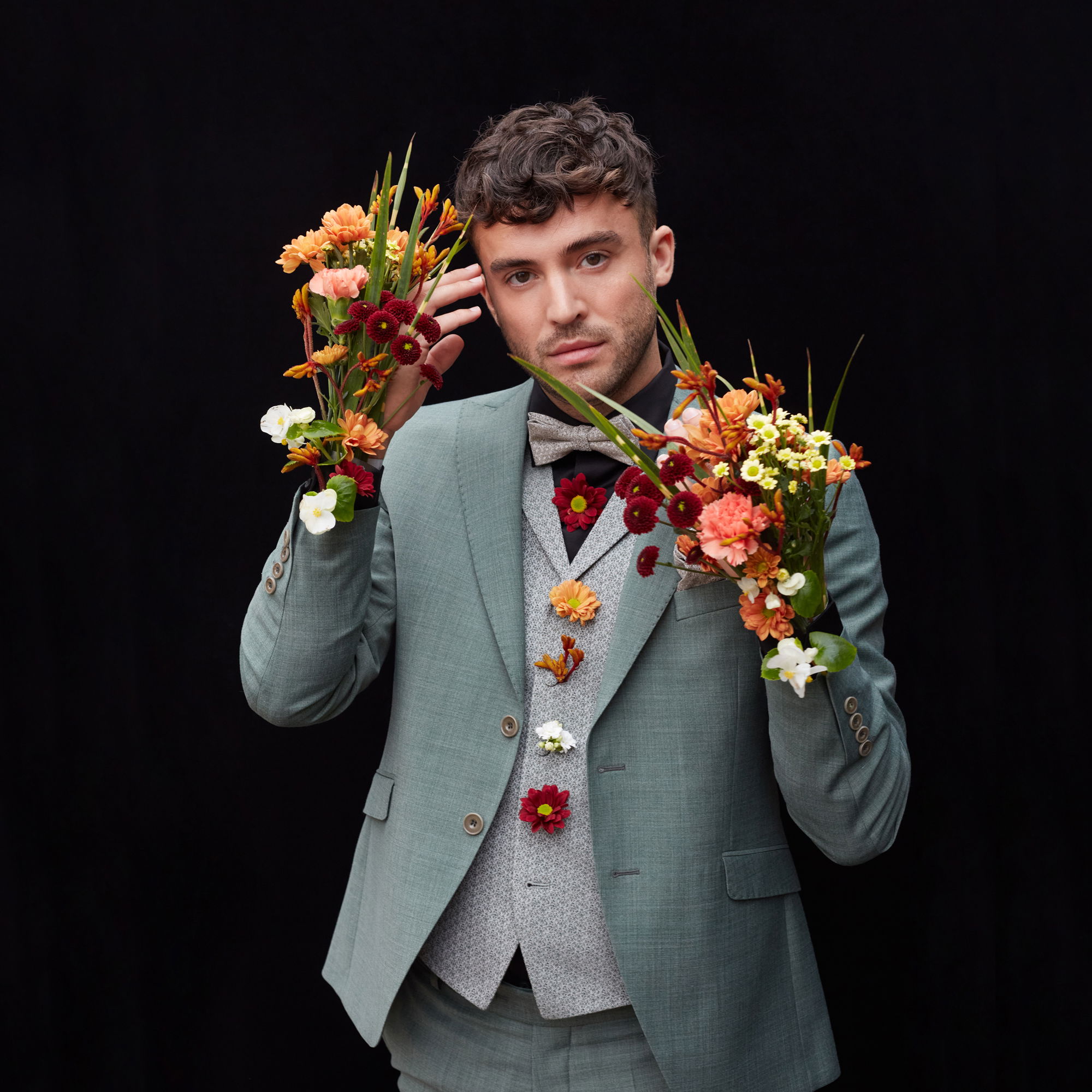 Ein junger Mann mit lockigen Haaren trägt einen hellgrünen Anzug und Accessoires aus Blumen in warmen Farben wie Orange, Rot und Gelb. Er steht vor einem dunklen Hintergrund.