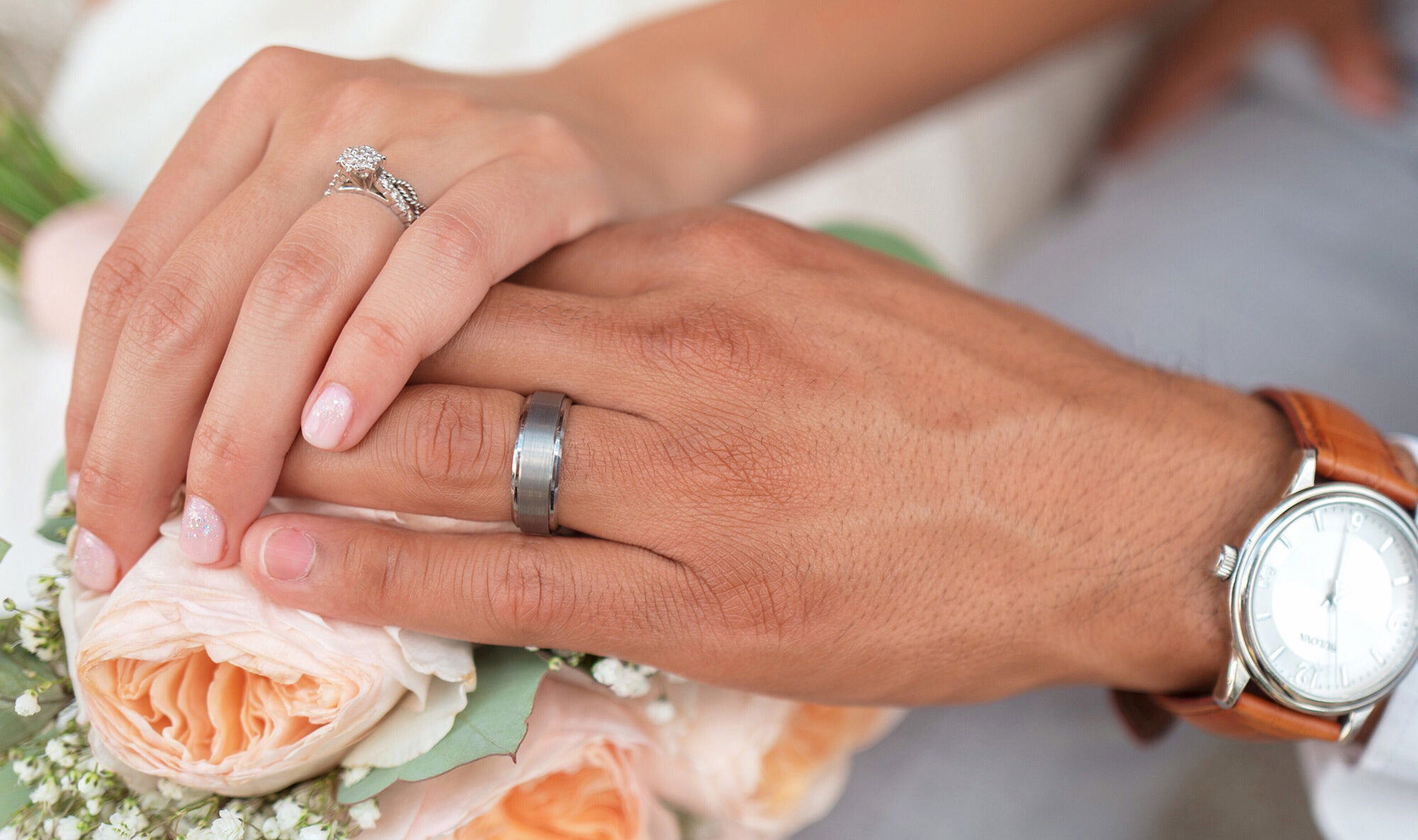 Ein Brautpaar hält sanft die Hände übereinander, um ihre Eheringe zu zeigen. Der Bräutigam trägt eine Uhr, während der Brautstrauß aus Rosen das Bild abrundet.
