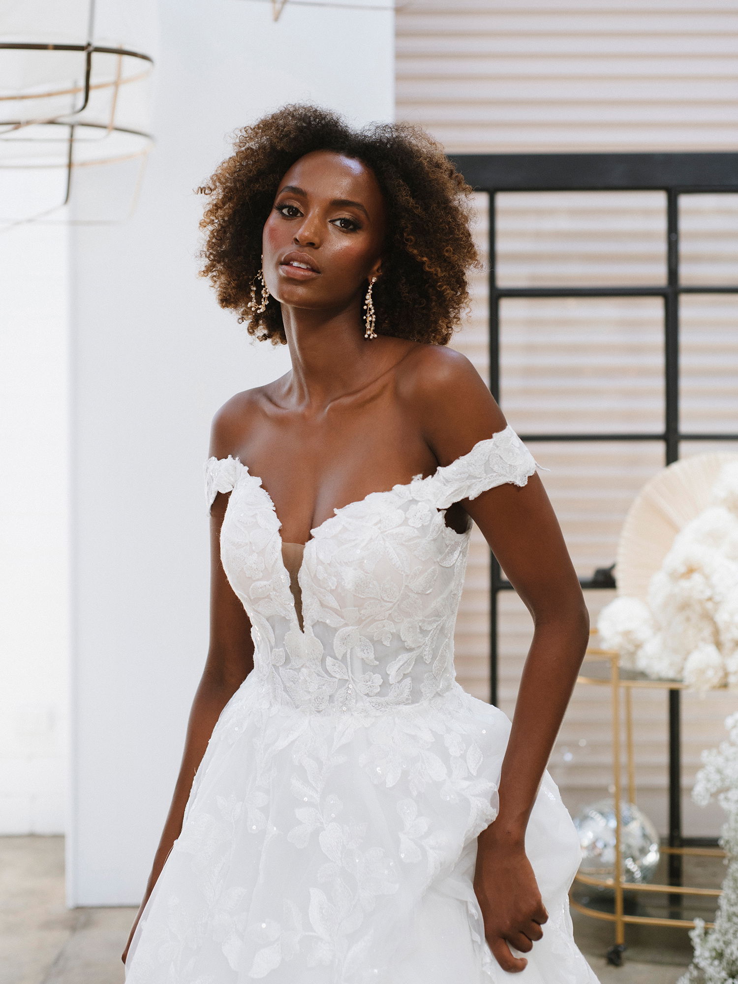 Frau in einem weißen, trägerlosen Kleid mit tiefem Ausschnitt und filigranen Blumendetails steht in einem stilvollen Raum und posiert mit einem eleganten Blick.