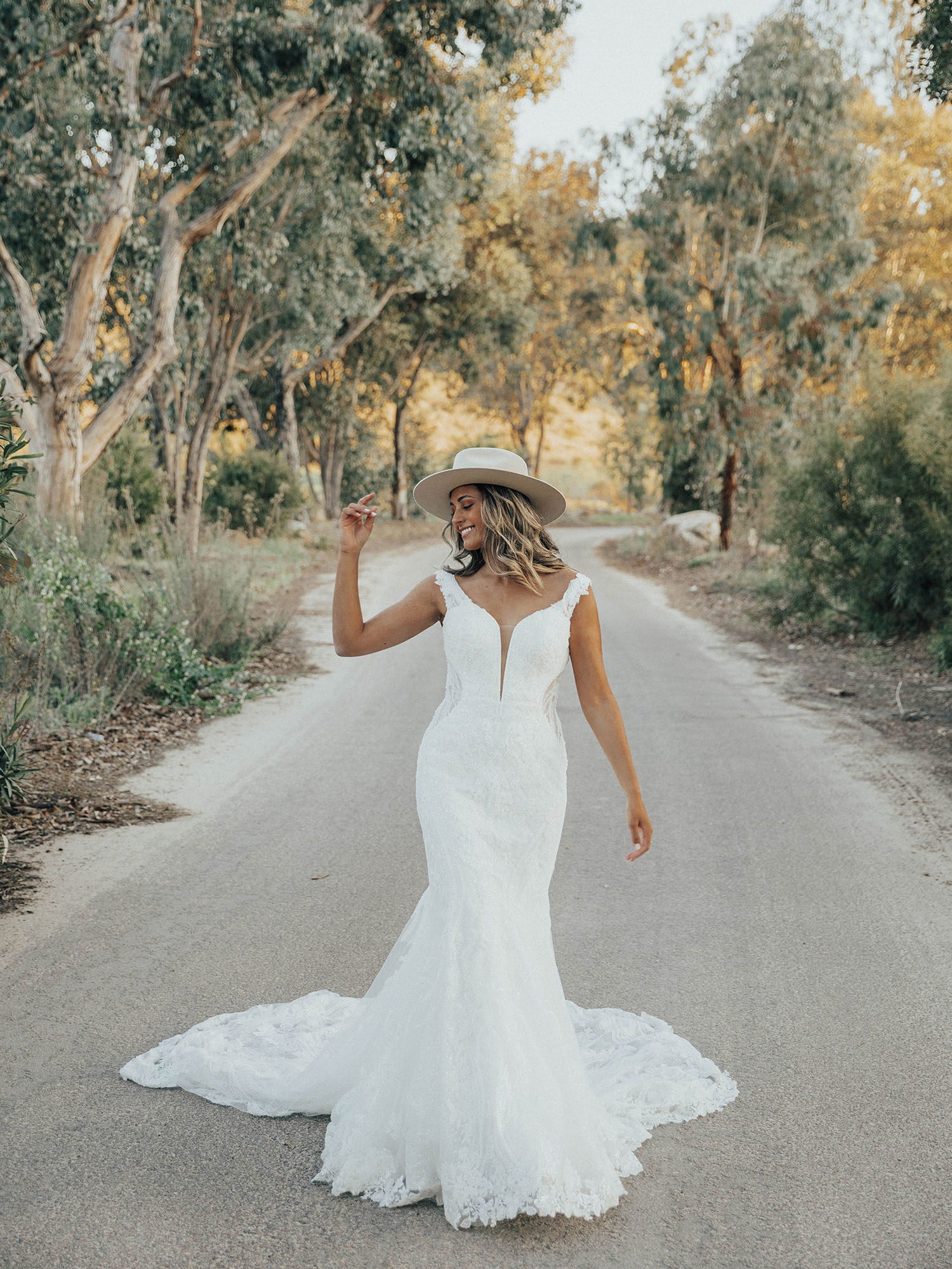 Braut in einem eleganten, figurschmeichelnden Hochzeitskleid mit einem breiten Hut auf dem Kopf, die entspannt auf einer Baumgesäumten Straße spaziert und dabei die Natur genießt.