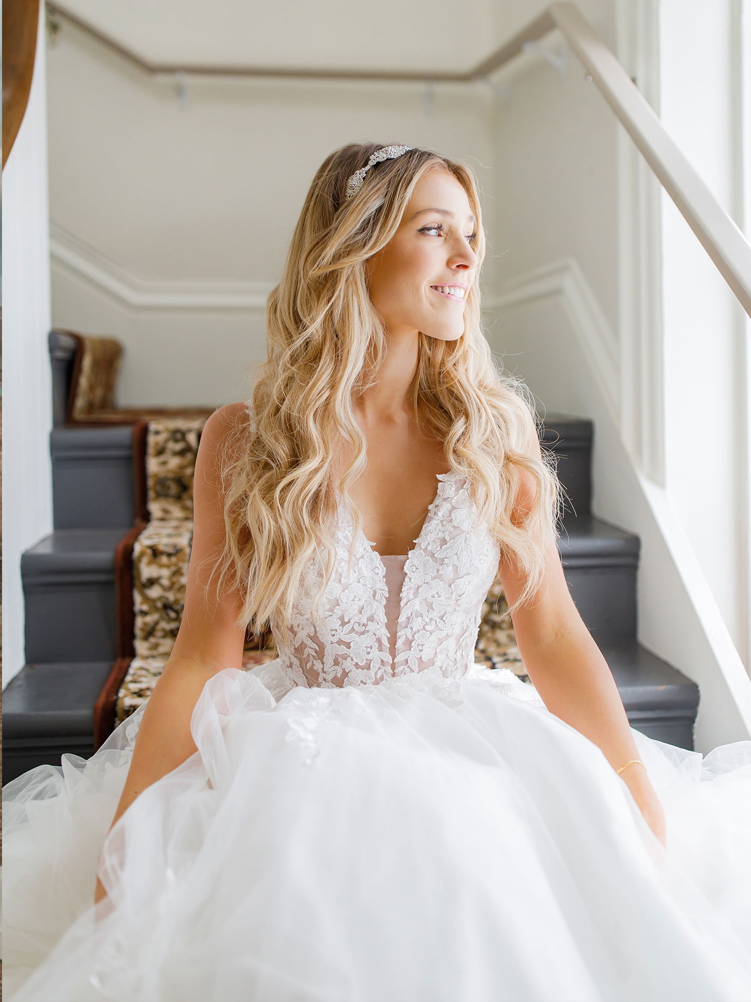 Frau in einem weißen, aufwendig verzierten Brautkleid sitzt lächelnd auf einer Treppe, das Kleid mit floralen Details und tiefem Ausschnitt erstreckt sich um sie herum.