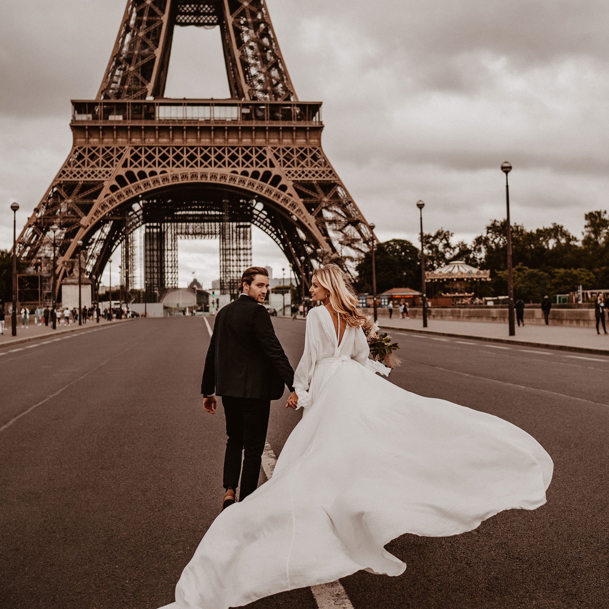 Das Brautpaar spaziert Hand in Hand vor dem Eiffelturm. Die Braut im weißen Kleid blickt lächelnd zurück, während ihr Schleier im Wind flattert.