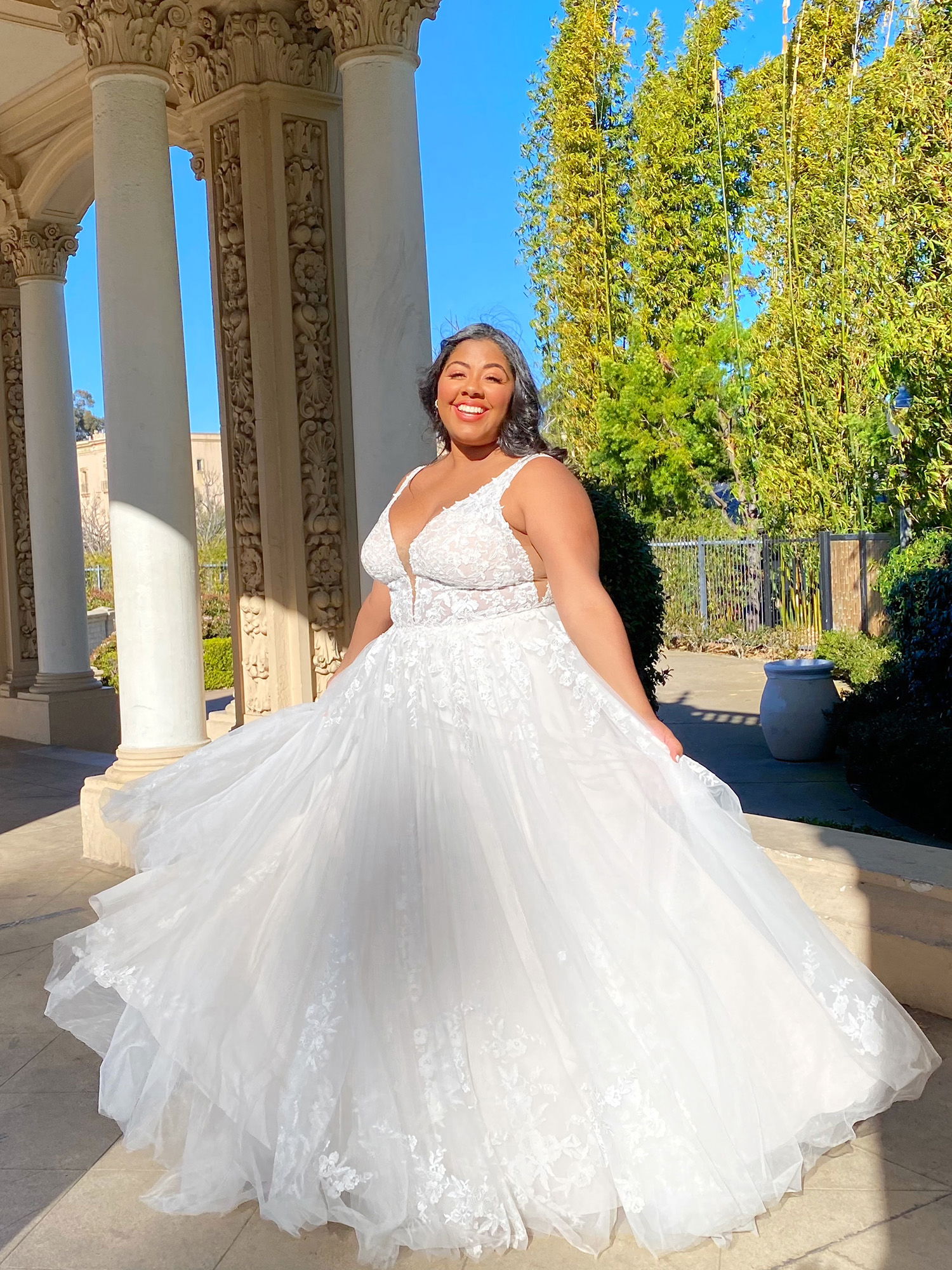 Braut in einem romantischen, weißen Brautkleid mit tiefem V-Ausschnitt und voluminösem Rock lächelt fröhlich, während sie in einem sonnigen Außenbereich unter dekorativen Säulen posiert.