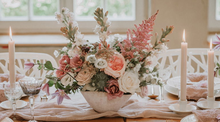 Eine romantische Blumendekoration mit rosa, weißen und lila Blumen, arrangiert in einer Schale, steht auf einem Hochzeitstisch, flankiert von brennenden Kerzen und feinem Geschirr.