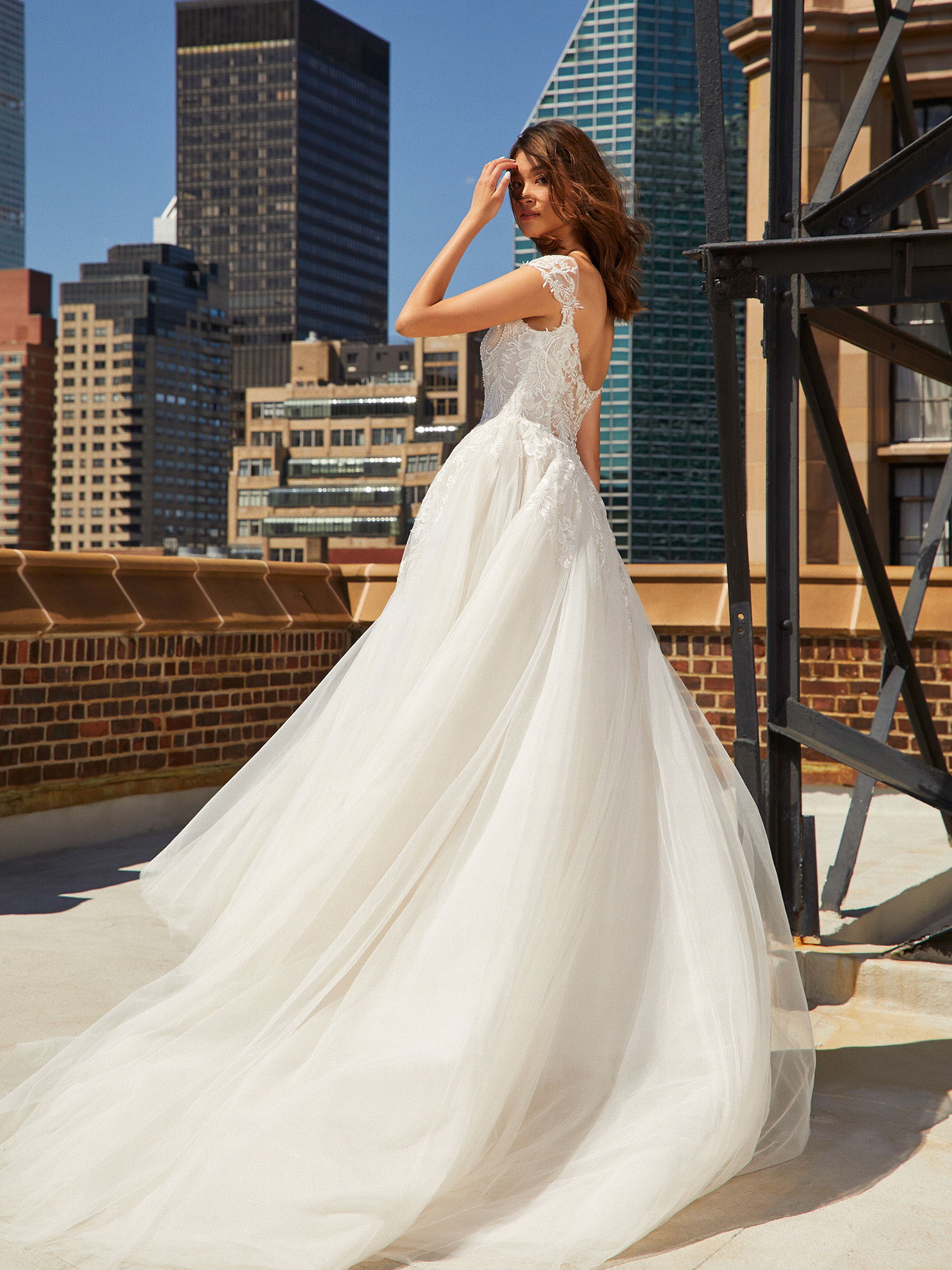 Die Braut in einem voluminösen, rückenfreien Brautkleid posiert auf einer Dachterrasse. Ihr Kleid mit Spitzenapplikationen und weitem Tüllrock wirkt besonders glamourös vor der urbanen Kulisse von Hochhäusern und modernen Gebäuden.