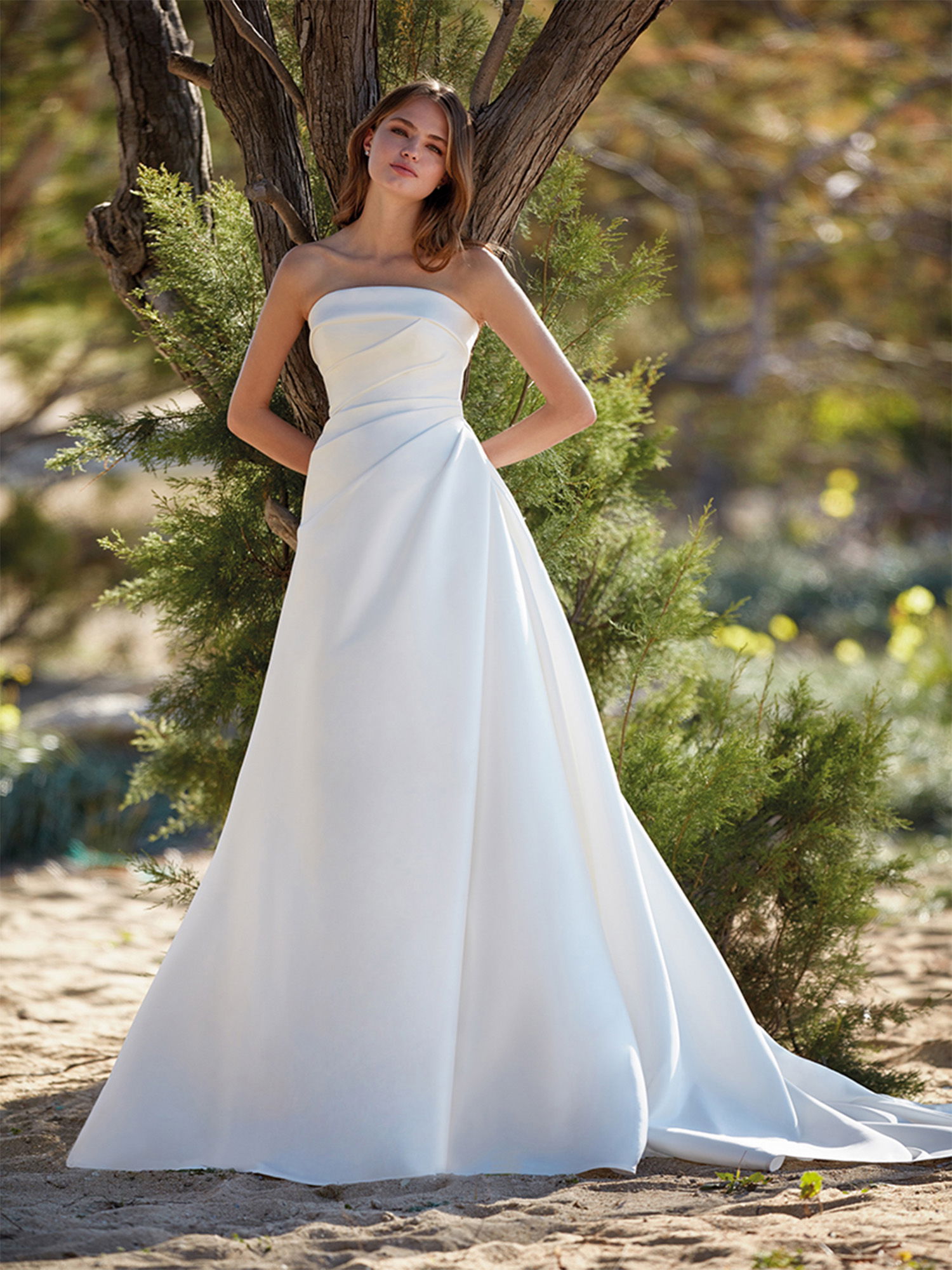 Die Braut steht in einem minimalistischen, trägerlosen Brautkleid mit weitem Rock vor einem Baum. Der Naturhintergrund, mit Büschen und Sand, verleiht der Szene eine ruhige und entspannte Atmosphäre.