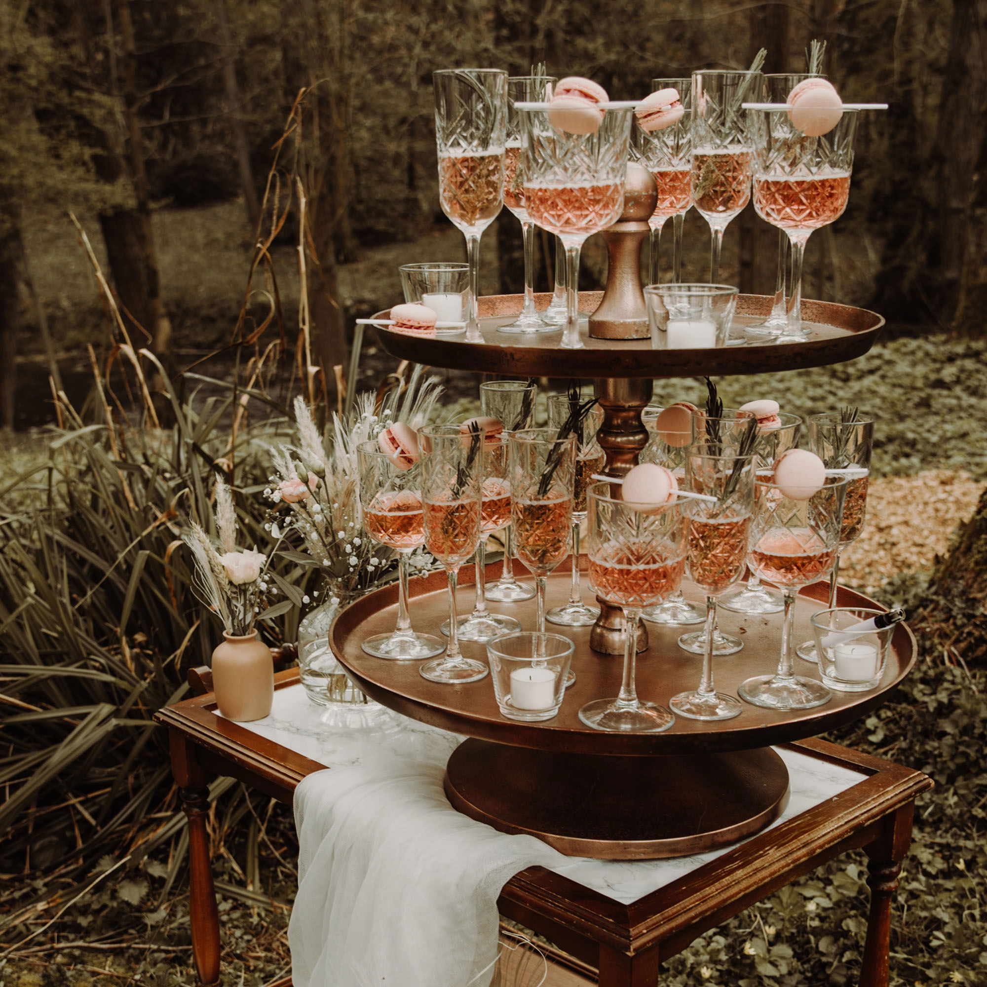 Doppelt gestapeltes Serviergestell mit Kristallgläsern, gefüllt mit Rosé-Champagner und dekoriert mit rosa Macarons. Das Arrangement steht auf einem rustikalen Holztisch, umgeben von einem grünen, bewaldeten Hintergrund, der der Hochzeitsfeier einen eleganten, naturverbundenen Touch gibt.