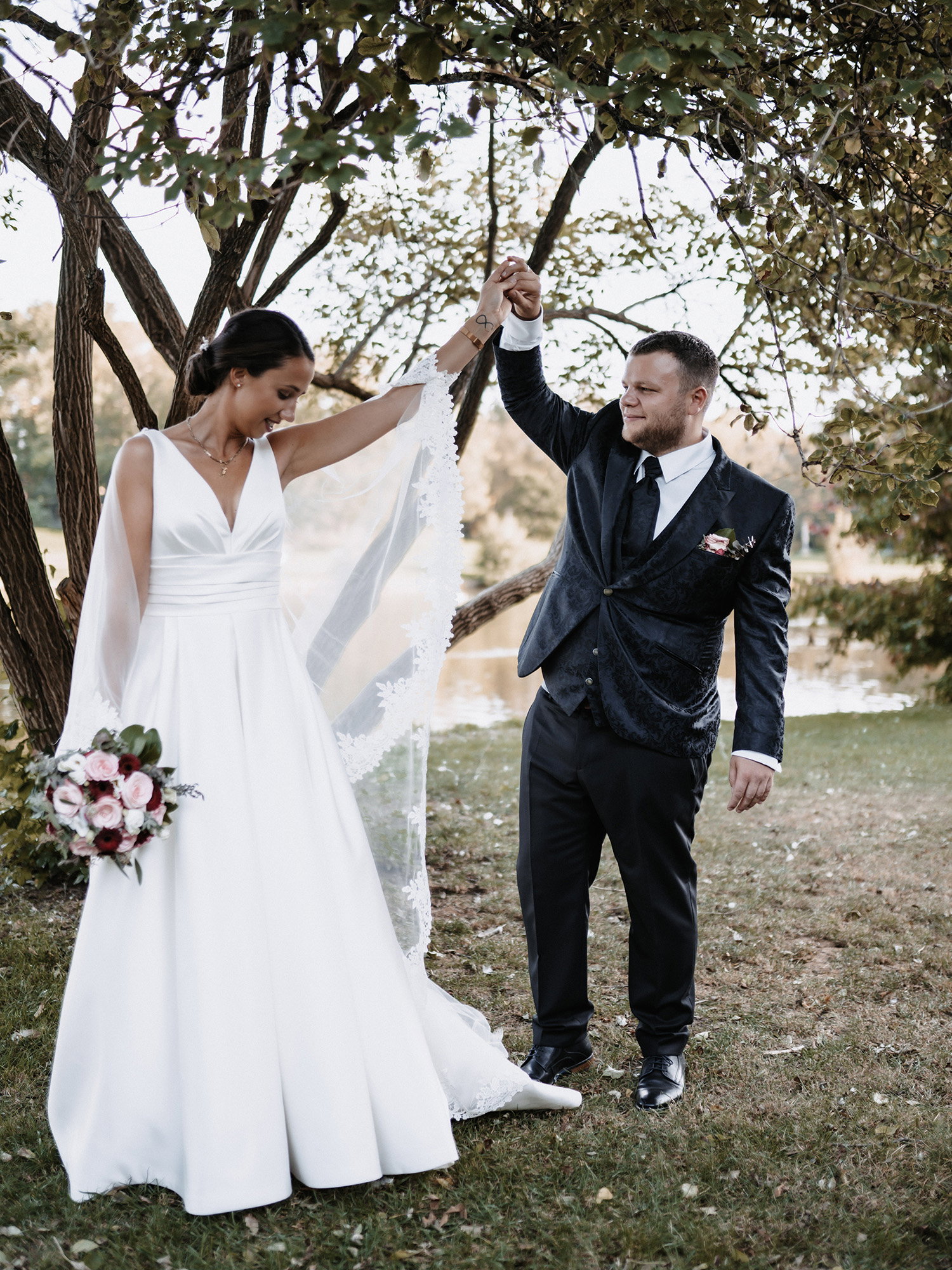 Das Brautpaar Claudia und Hendrik tanzt unter einem Baum in der Natur. Die Braut hebt eine Hand, die mit dem Schleier geschmückt ist, während sie einen floralen Brautstrauß in der anderen Hand hält. Der Bräutigam in einem dunklen Anzug hebt ihre Hand, als ob sie zu einem Tanzschritt ansetzen. Die natürliche Umgebung am See bildet den Hintergrund dieser zarten Szene.
